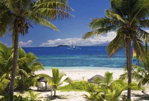 "Tropical beach with palm trees, white sand, and sailboats on turquoise ocean under blue sky"