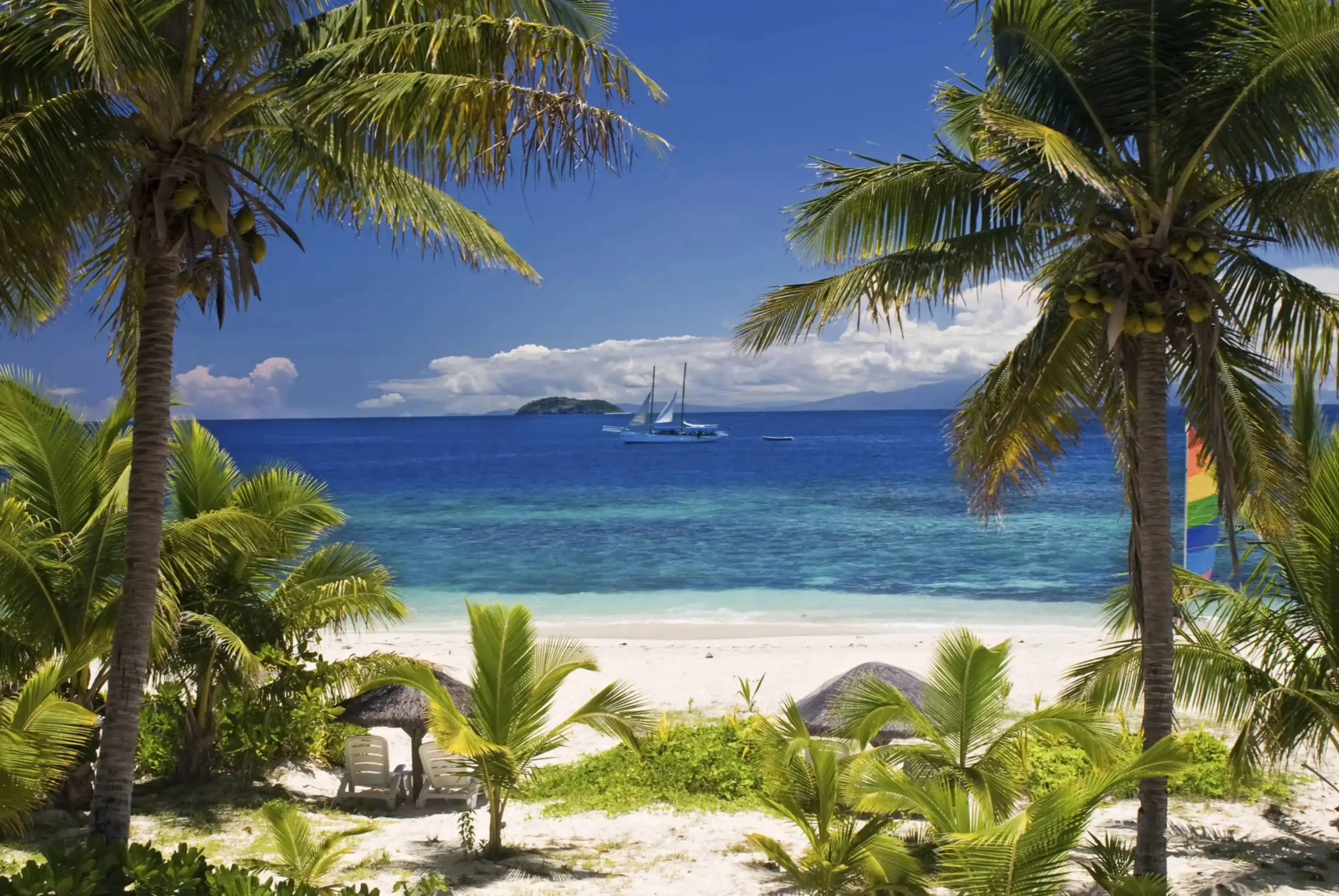 "Tropical beach with palm trees, white sand, and sailboats on turquoise ocean under blue sky"