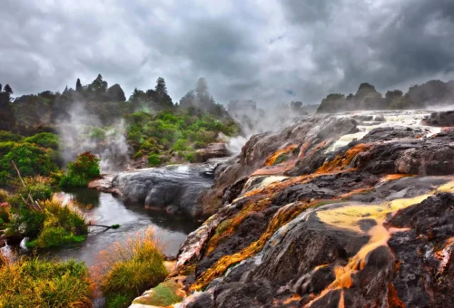 "Steaming volcanic terrain with flowing lava and lush greenery under cloudy sky"
