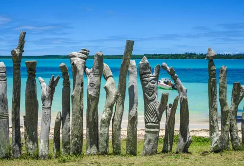 "Traditional carved wooden totems lined up on grassy shore with turquoise ocean and blue sky in the background"