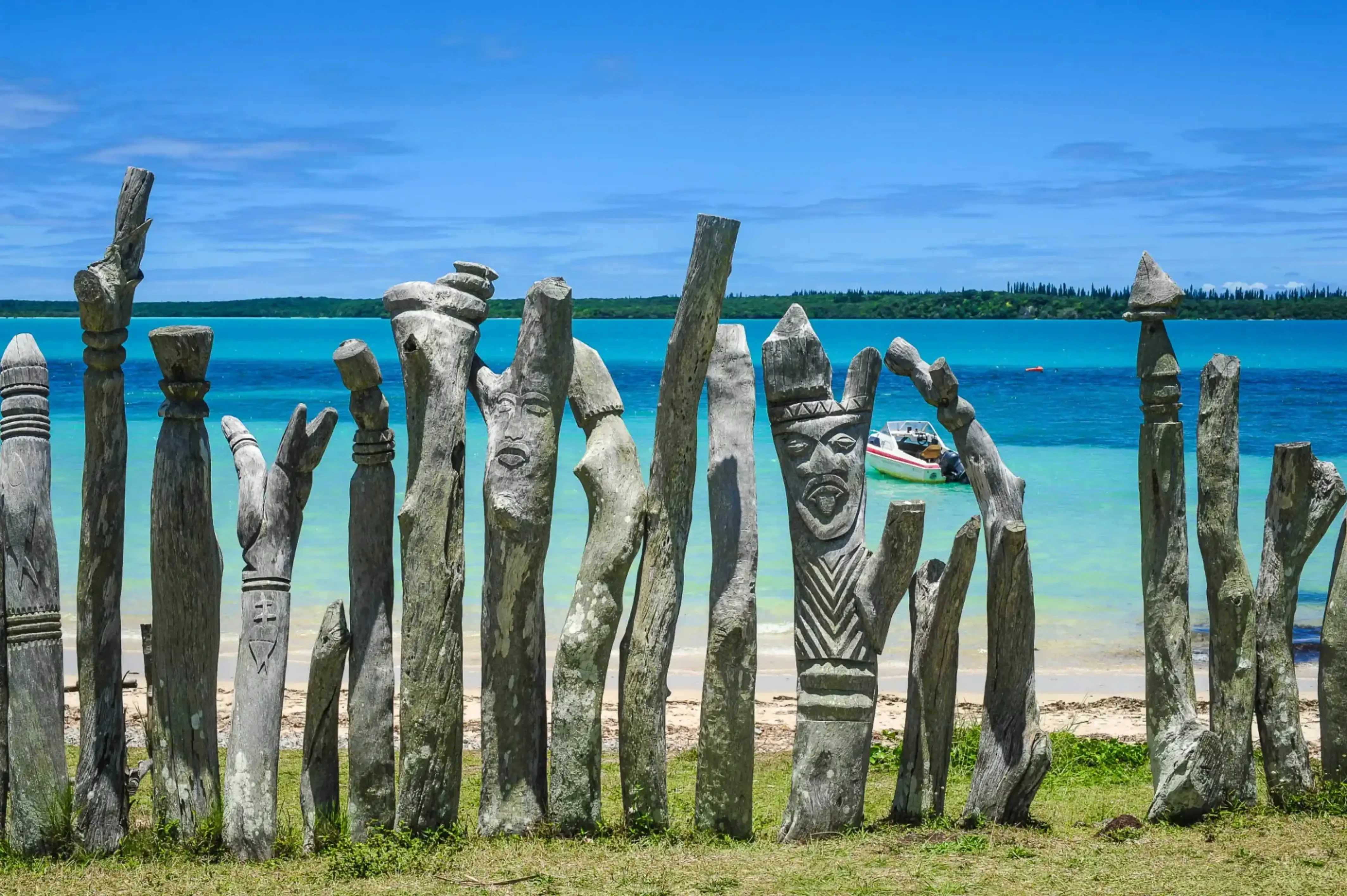 "Traditional carved wooden totems lined up on grassy shore with turquoise ocean and blue sky in the background"