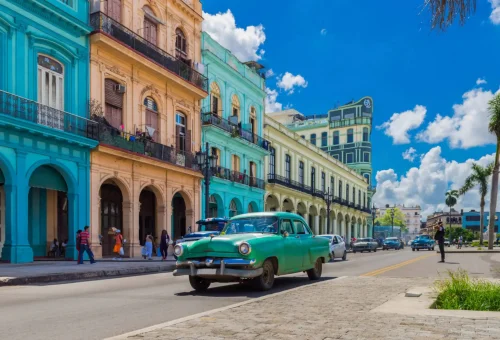 "Vintage green car driving past colorful colonial-style buildings under a bright blue sky"