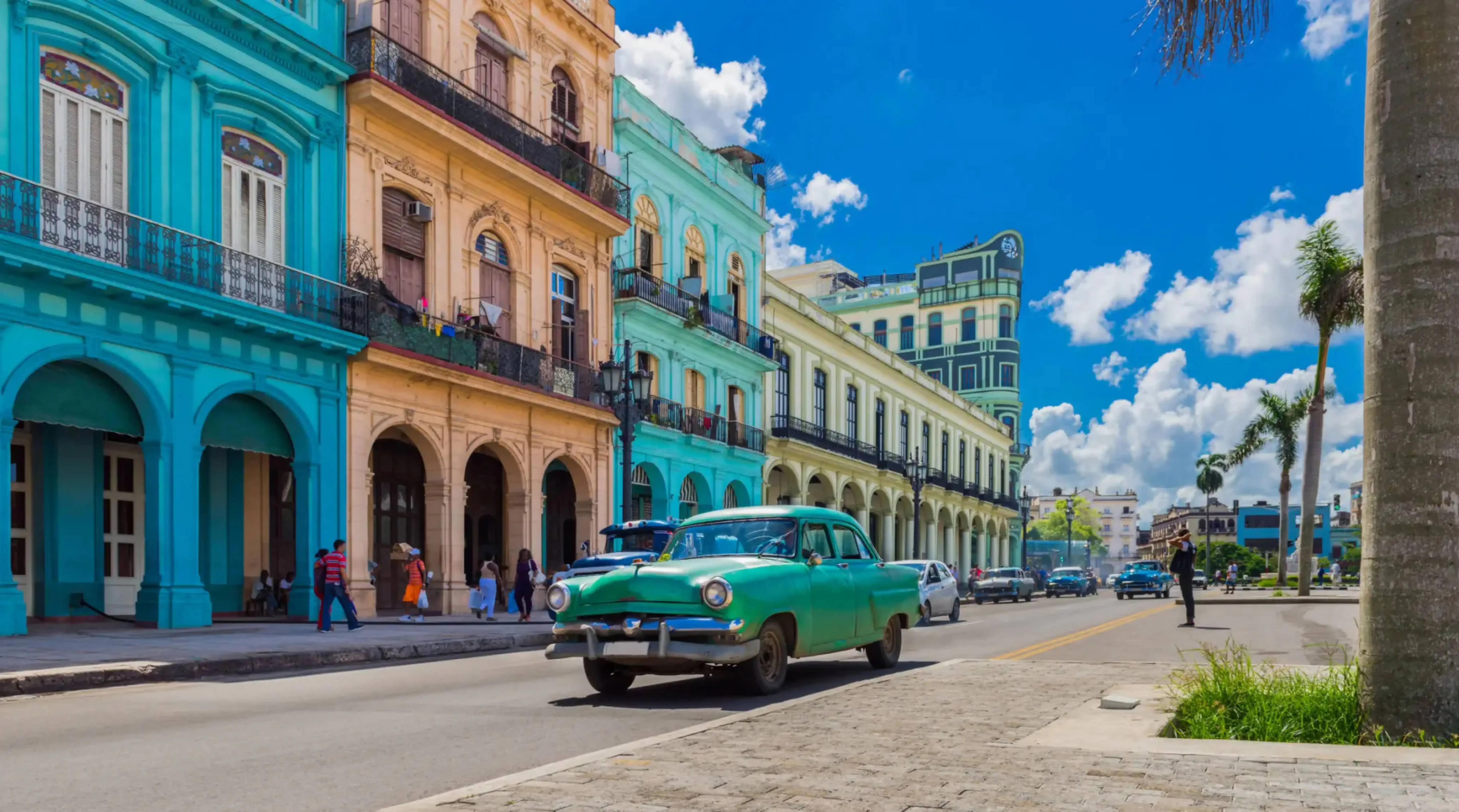 "Vintage green car driving past colorful colonial-style buildings under a bright blue sky"