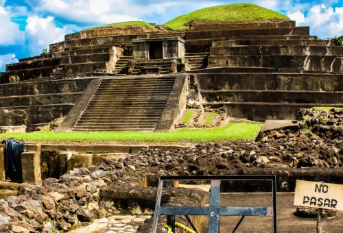 "Ancient terraced pyramid of the Tazumal ruins in Chalchuapa, El Salvador, with a 'No Pasar' sign in the foreground under a bright blue sky."