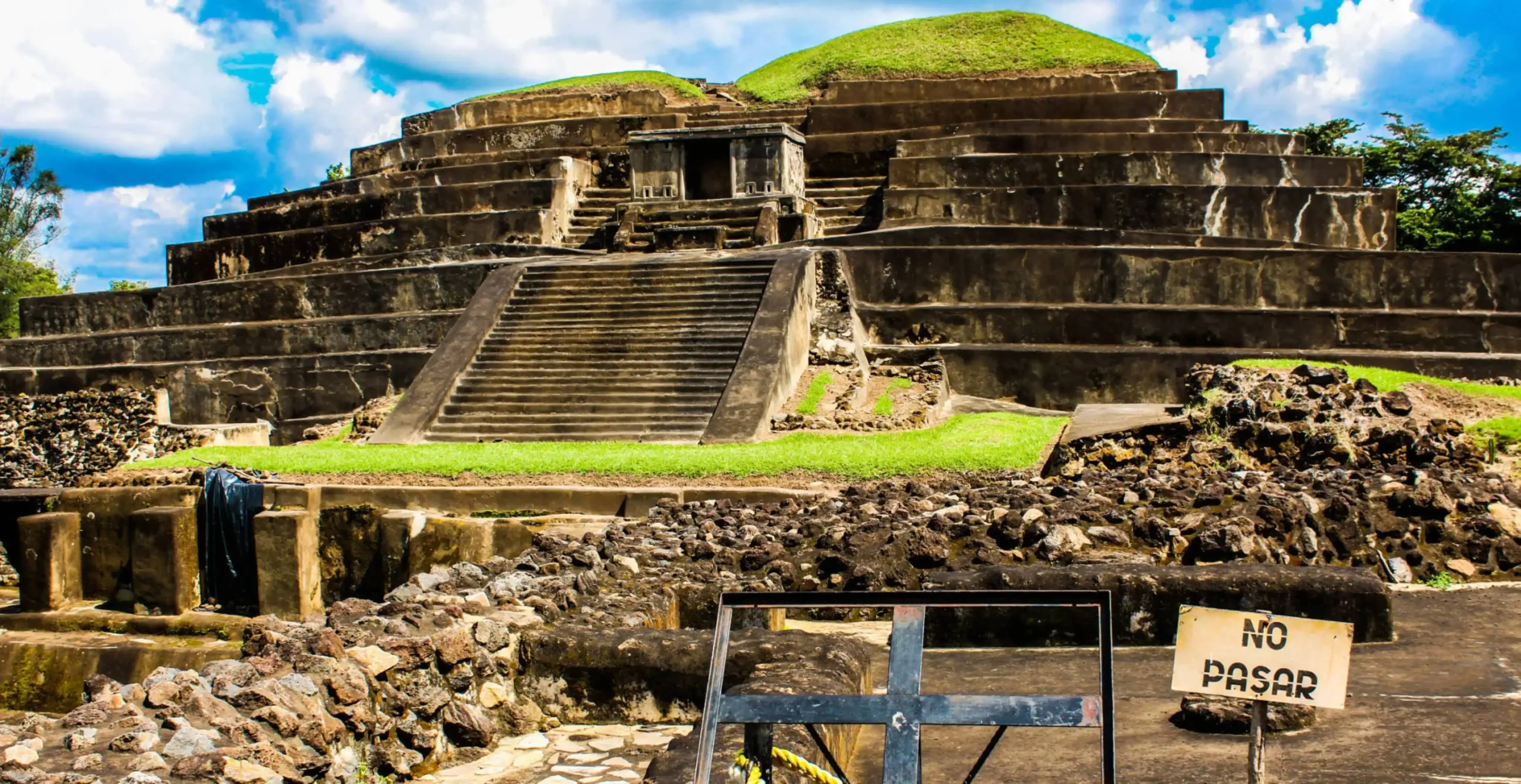 "Ancient terraced pyramid of the Tazumal ruins in Chalchuapa, El Salvador, with a 'No Pasar' sign in the foreground under a bright blue sky."