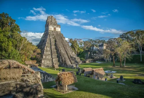 "Temple I of the ancient Mayan city of Tikal in Petén, Guatemala, with thatched huts and stone ruins under a clear blue sky."