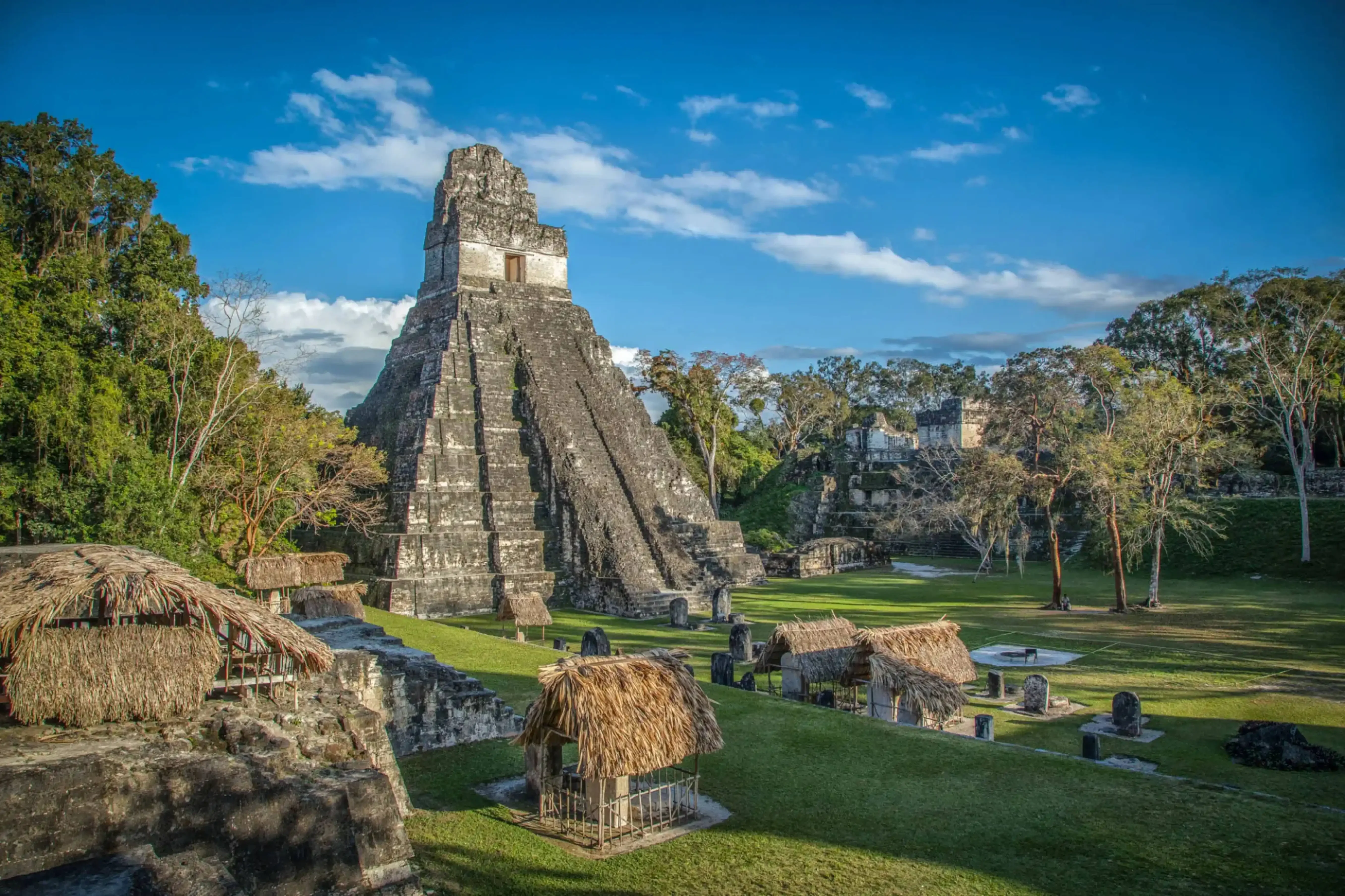 "Temple I of the ancient Mayan city of Tikal in Petén, Guatemala, with thatched huts and stone ruins under a clear blue sky."