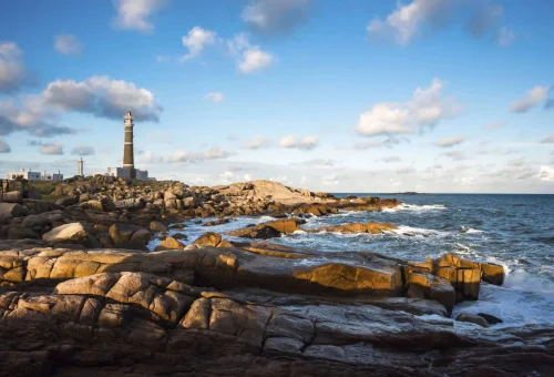 "Rocky coastline with waves crashing and the Punta del Este Lighthouse in the distance under a blue sky, Uruguay."