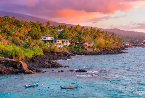 "Colorful sunset over a tropical coastal village with palm trees, fishing boats, and volcanic rock shoreline, possibly São Tomé and Príncipe."