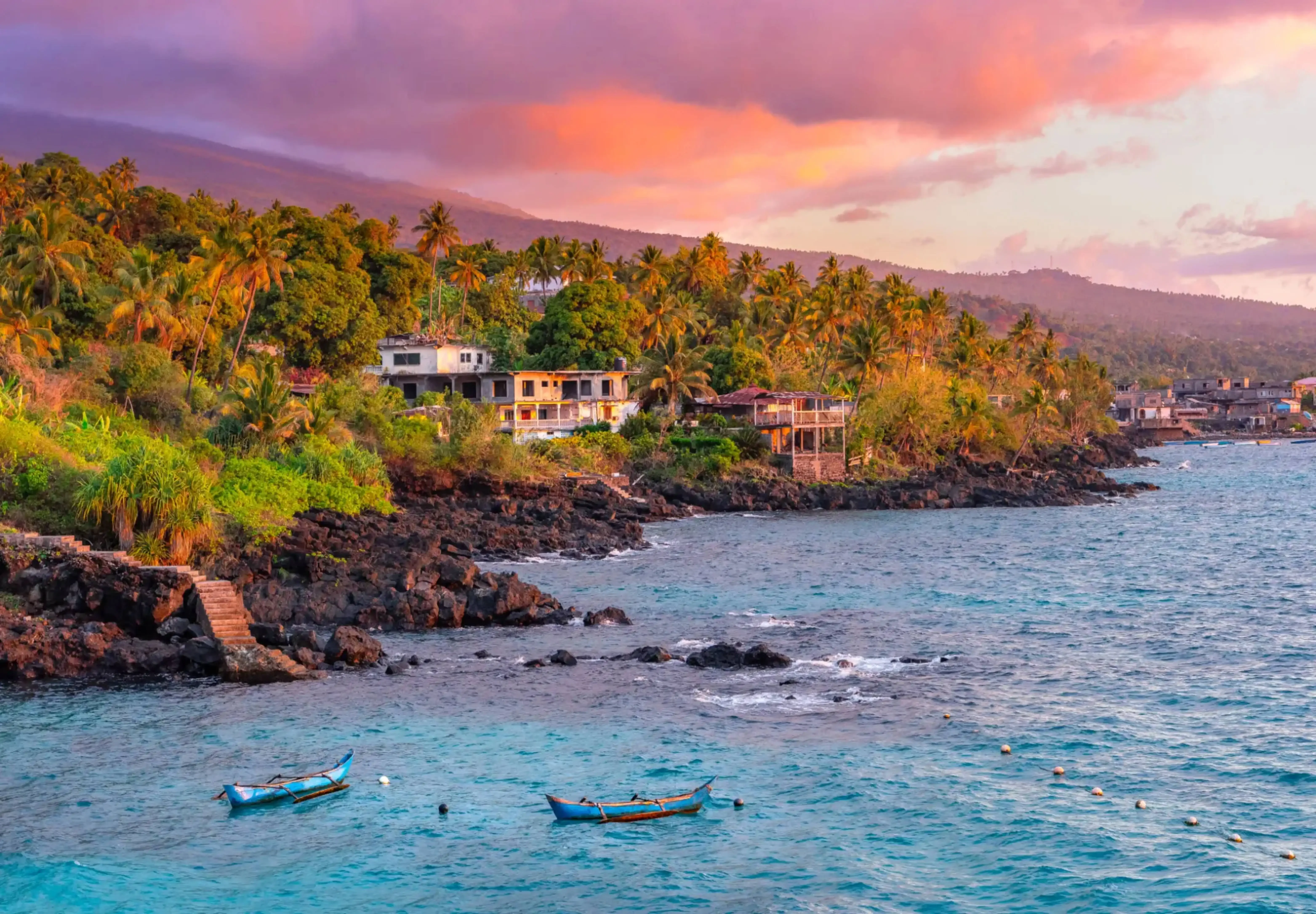 "Colorful sunset over a tropical coastal village with palm trees, fishing boats, and volcanic rock shoreline, possibly São Tomé and Príncipe."