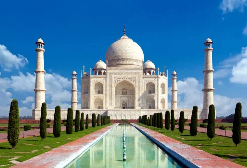 The Taj Mahal with its reflection in the central water channel, framed by manicured gardens and a bright blue sky.