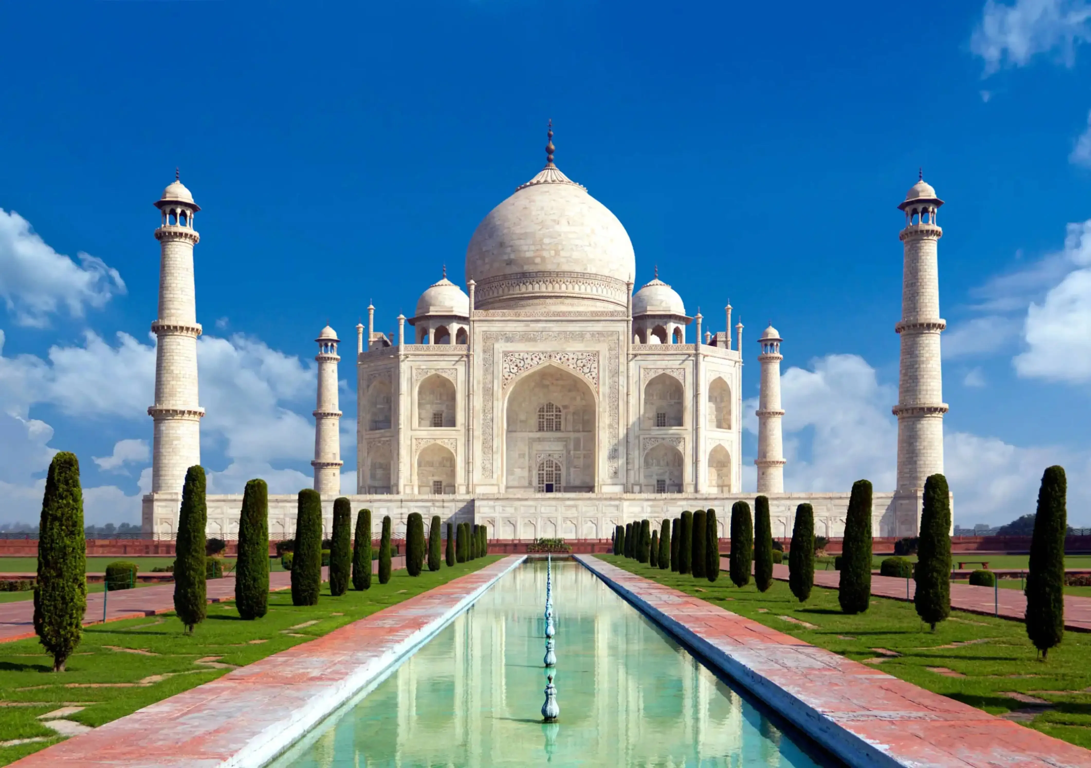 The Taj Mahal with its reflection in the central water channel, framed by manicured gardens and a bright blue sky.