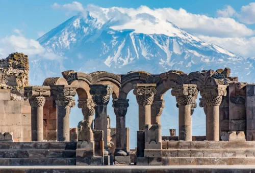 Ancient stone columns of the Zvartnots Cathedral ruins with snow-capped Mount Ararat rising majestically in the background.