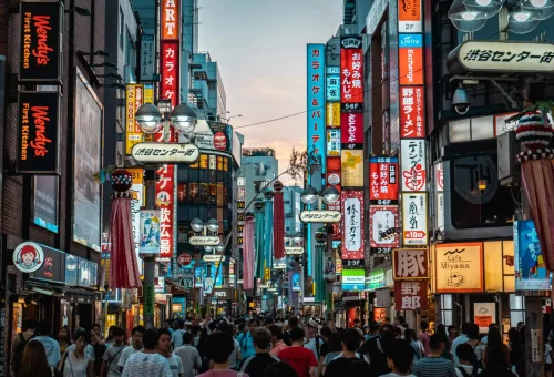 Crowded street scene in Shibuya, Tokyo, Japan, with vibrant neon signs and storefronts at dusk.