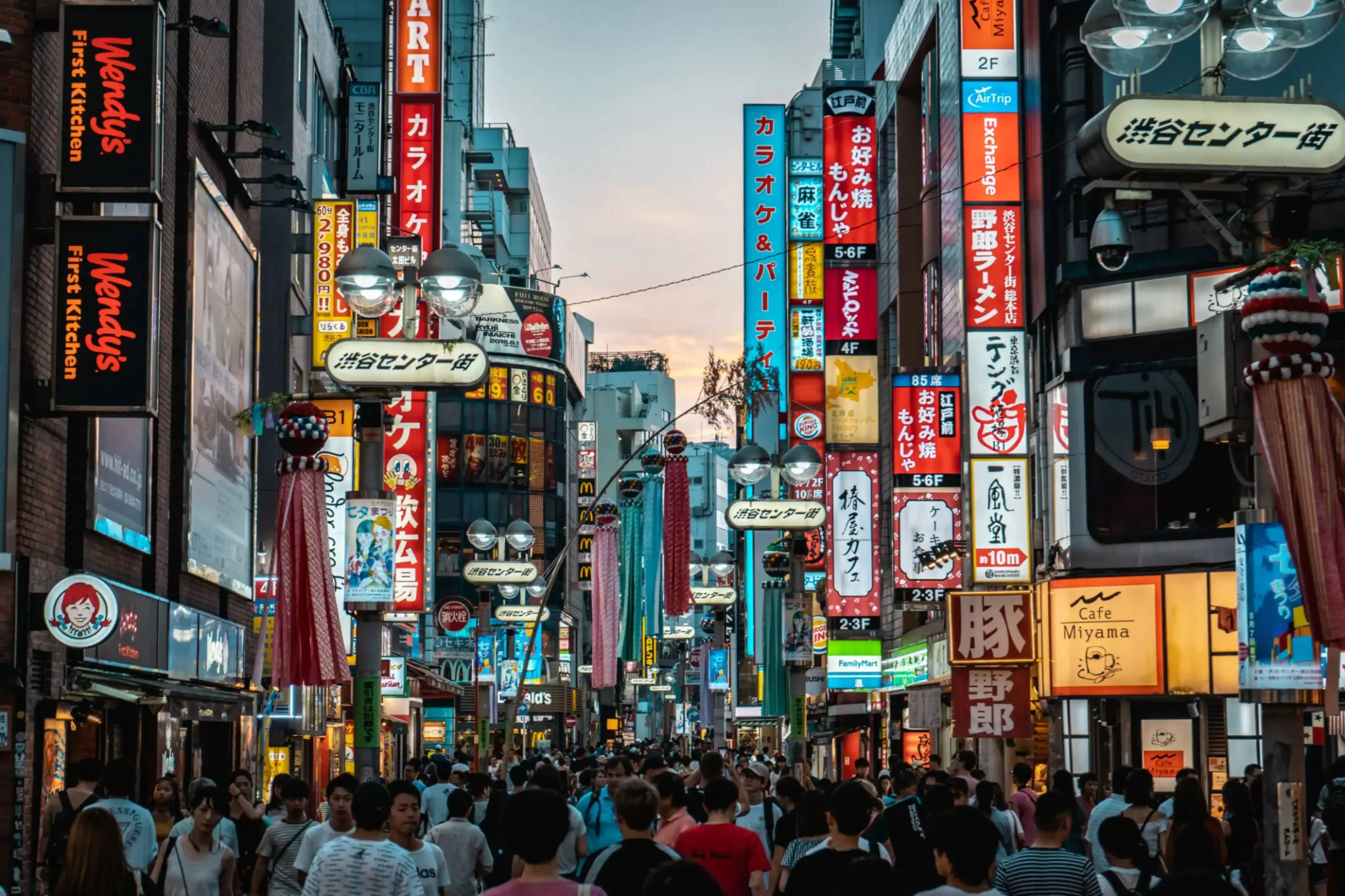 Crowded street scene in Shibuya, Tokyo, Japan, with vibrant neon signs and storefronts at dusk.