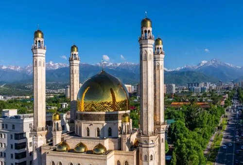Aerial view of the Central Mosque in Almaty, Kazakhstan, with golden domes and minarets set against the backdrop of the snow-capped Tian Shan mountains.