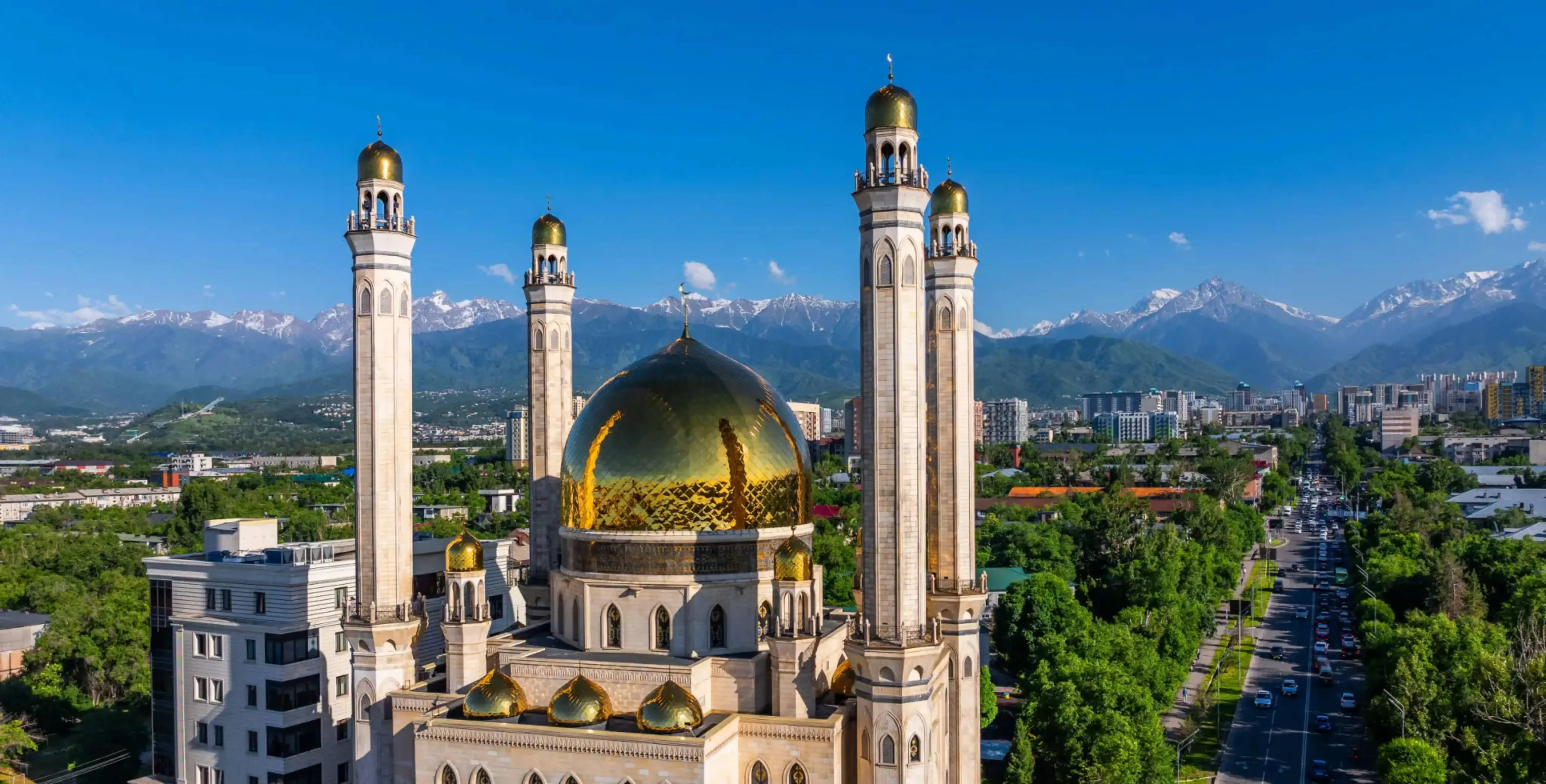 Aerial view of the Central Mosque in Almaty, Kazakhstan, with golden domes and minarets set against the backdrop of the snow-capped Tian Shan mountains.
