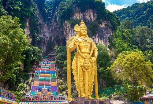 Golden statue of Lord Murugan at the entrance of Batu Caves in Malaysia, with colorful stairway leading into the limestone cave temple.
