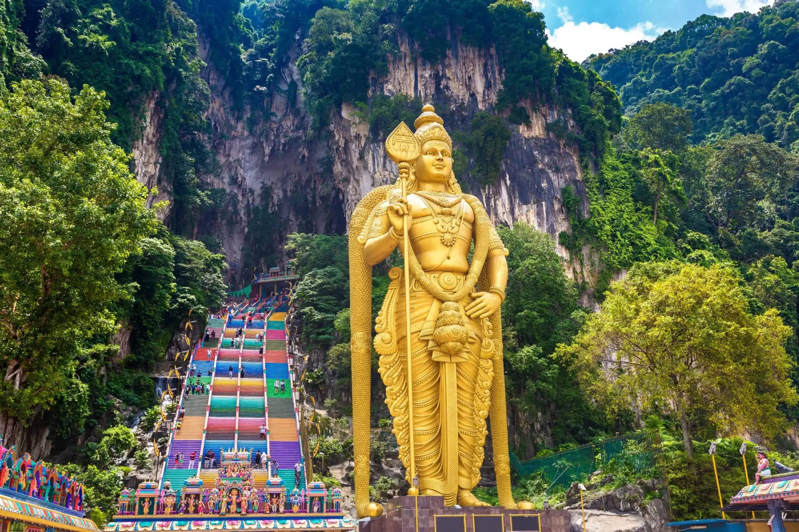 Golden statue of Lord Murugan at the entrance of Batu Caves in Malaysia, with colorful stairway leading into the limestone cave temple.