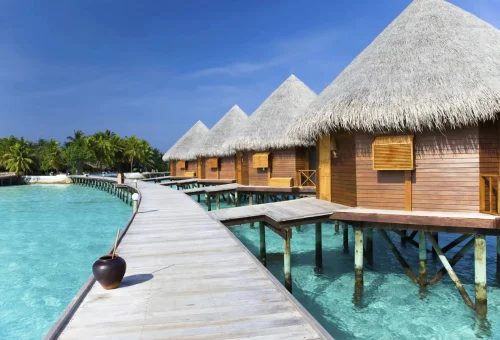 Wooden overwater bungalows with thatched roofs in the Maldives, connected by a boardwalk over crystal-clear turquoise lagoon.