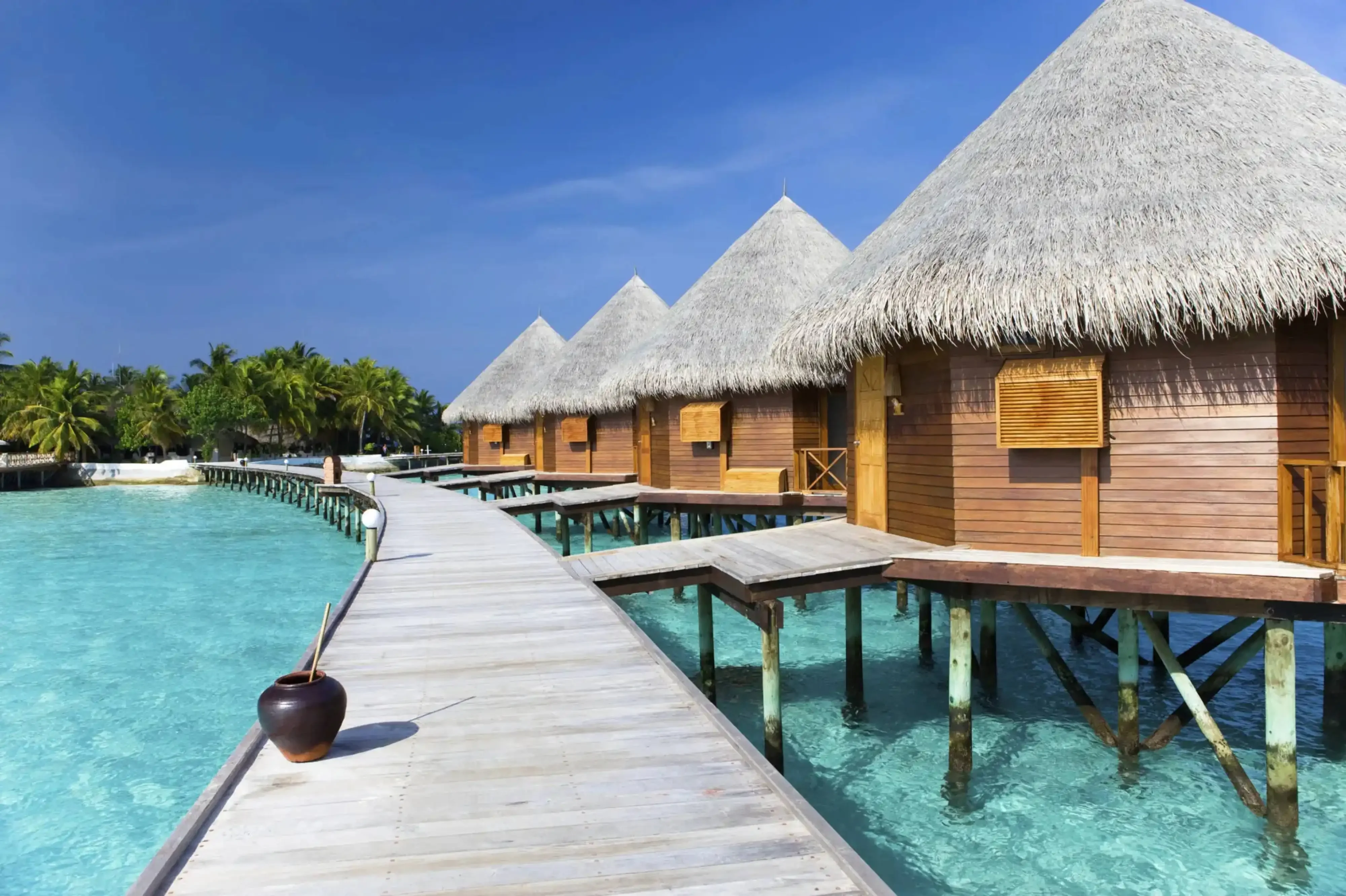Wooden overwater bungalows with thatched roofs in the Maldives, connected by a boardwalk over crystal-clear turquoise lagoon.