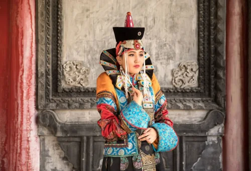Woman dressed in traditional Mongolian attire with elaborate headgear and colorful embroidered garments, standing against an ornate stone backdrop.