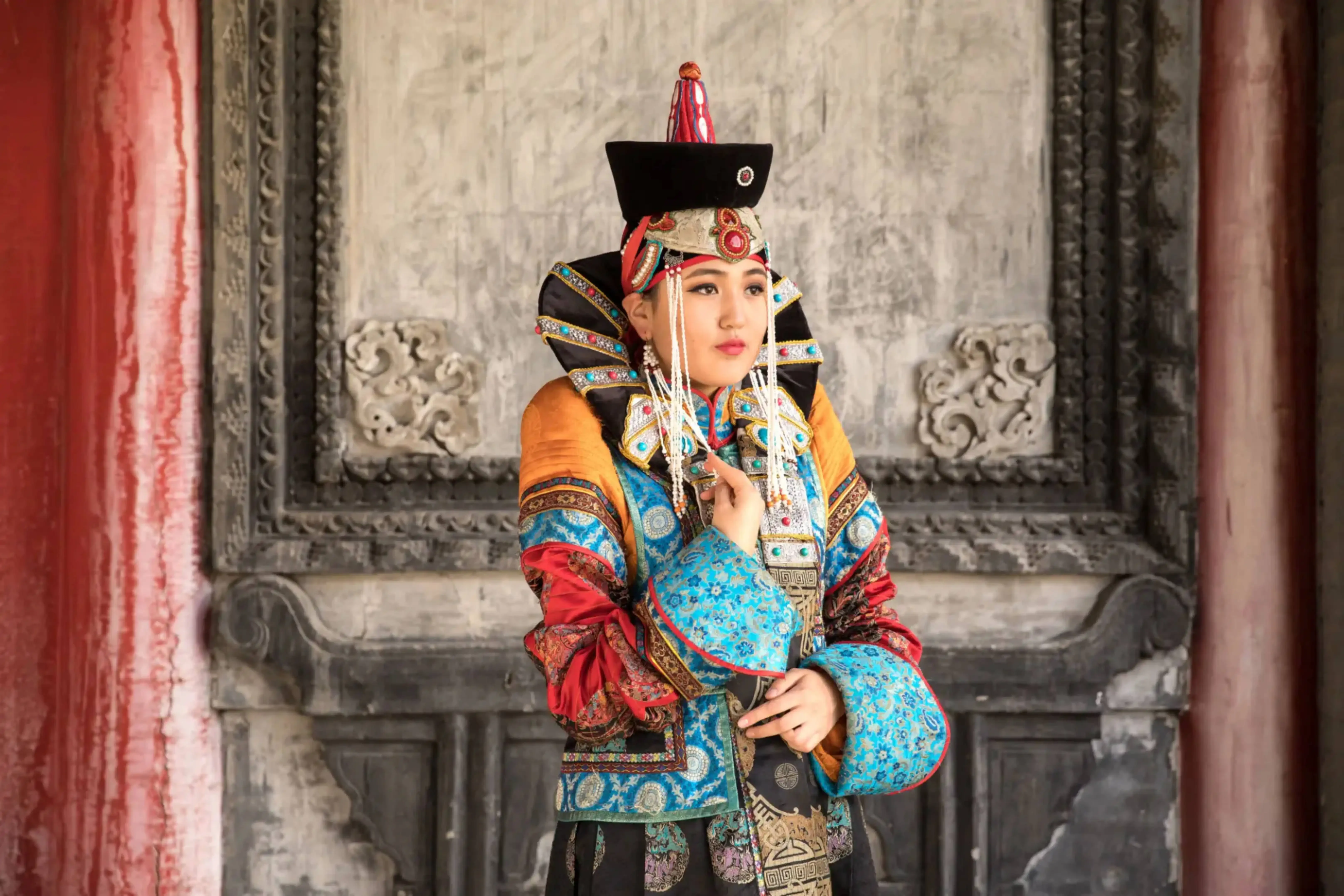 Woman dressed in traditional Mongolian attire with elaborate headgear and colorful embroidered garments, standing against an ornate stone backdrop.