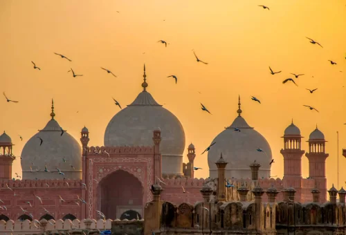 Birds flying over the Badshahi Mosque at sunset in Lahore, Pakistan, with domes and minarets silhouetted against a golden sky.