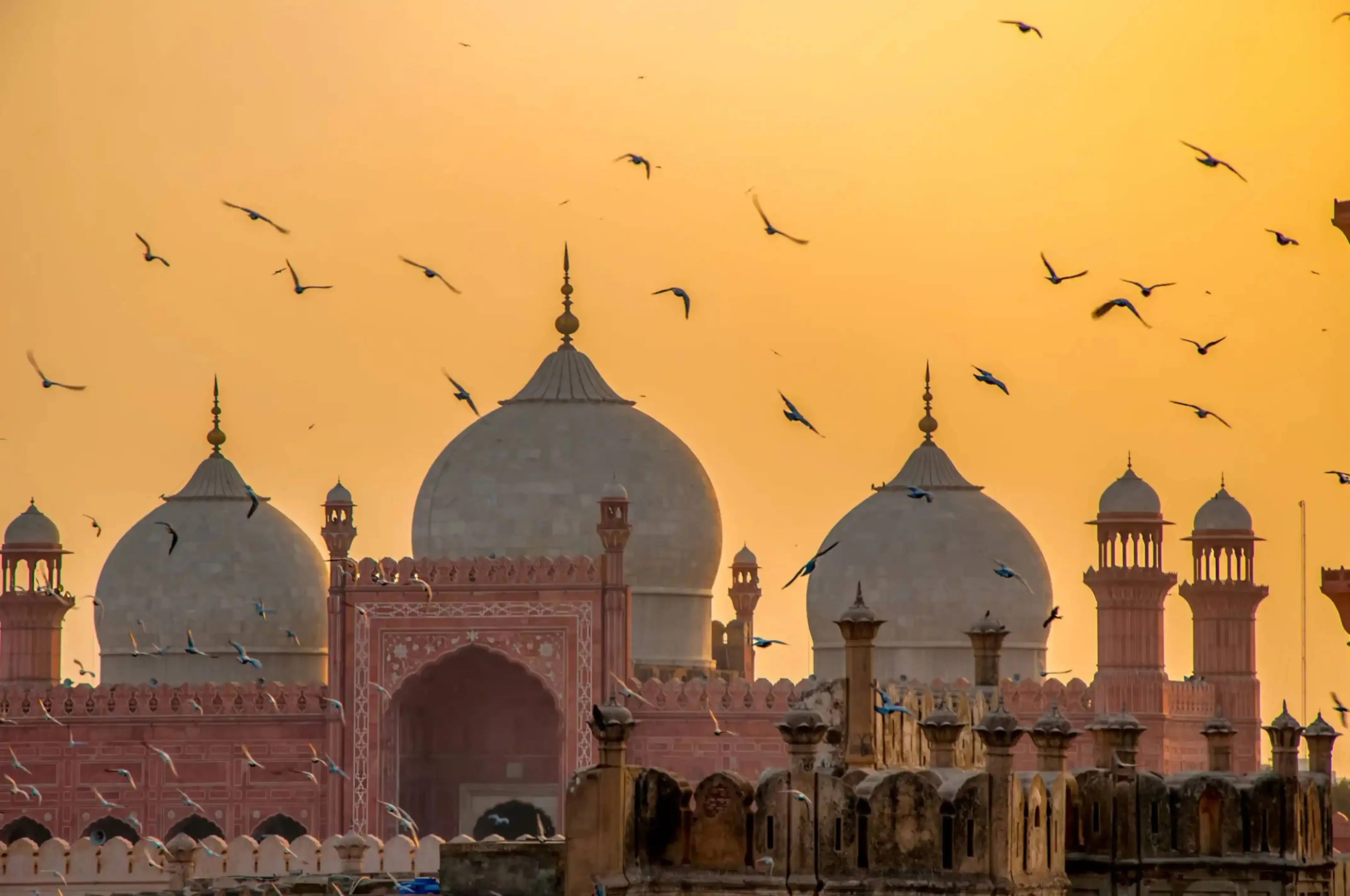 Birds flying over the Badshahi Mosque at sunset in Lahore, Pakistan, with domes and minarets silhouetted against a golden sky.