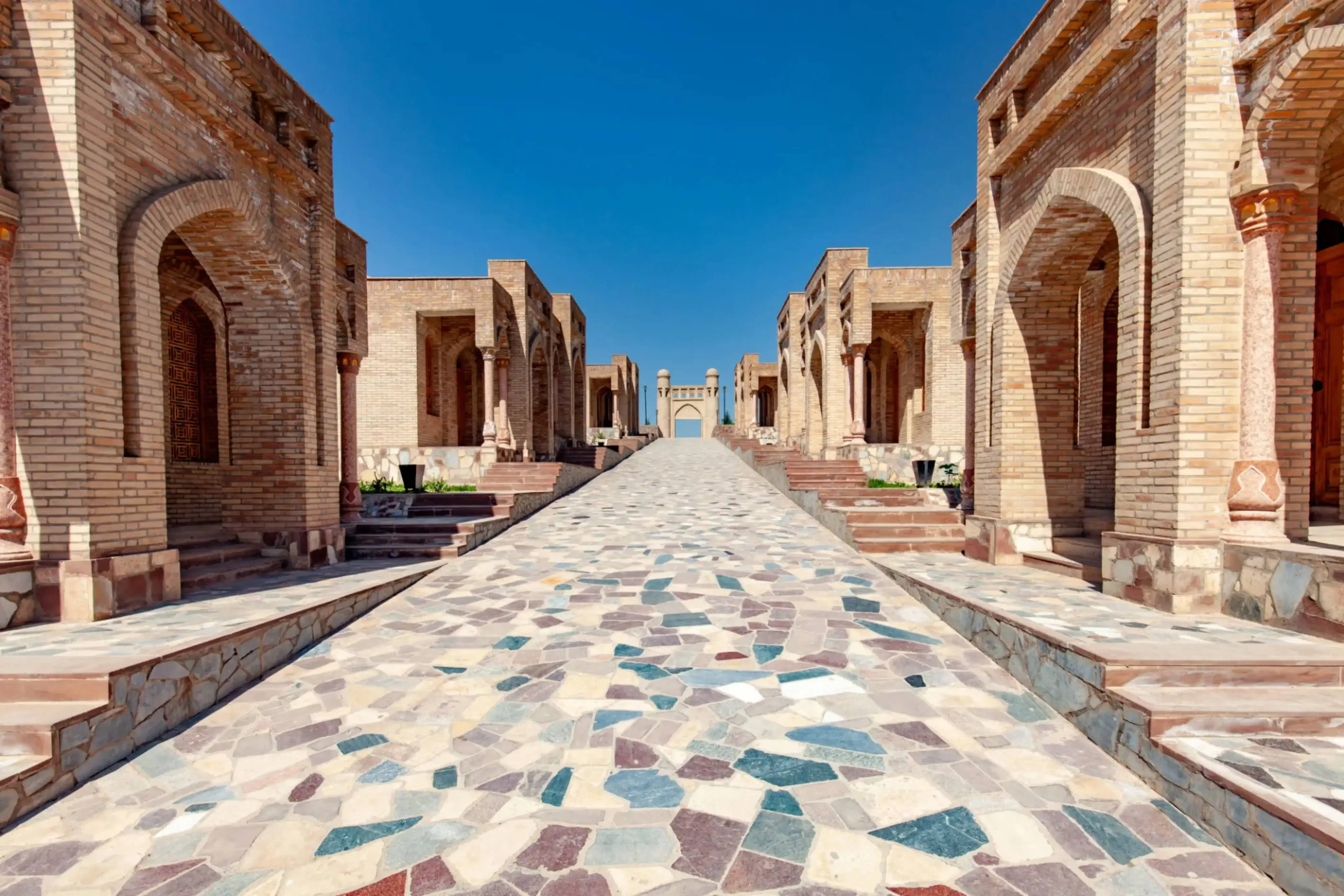 View of the Shah-i-Zinda necropolis in Samarkand, Uzbekistan, featuring a stone mosaic walkway lined with historic mausoleums and intricate Islamic architecture under a clear blue sky.