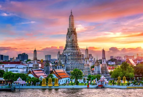 Wat Arun (Temple of Dawn) in Bangkok, Thailand, standing tall on the west bank of the Chao Phraya River with intricate spires and a glowing sunset sky in the background.