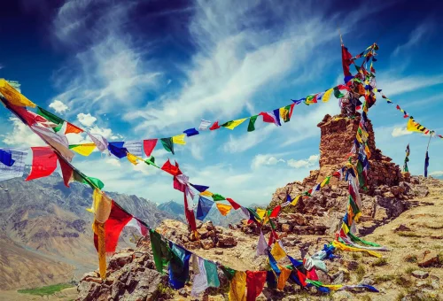 Colorful Tibetan prayer flags fluttering on a rocky mountain pass in the Himalayas, set against a dramatic blue sky with wispy clouds.