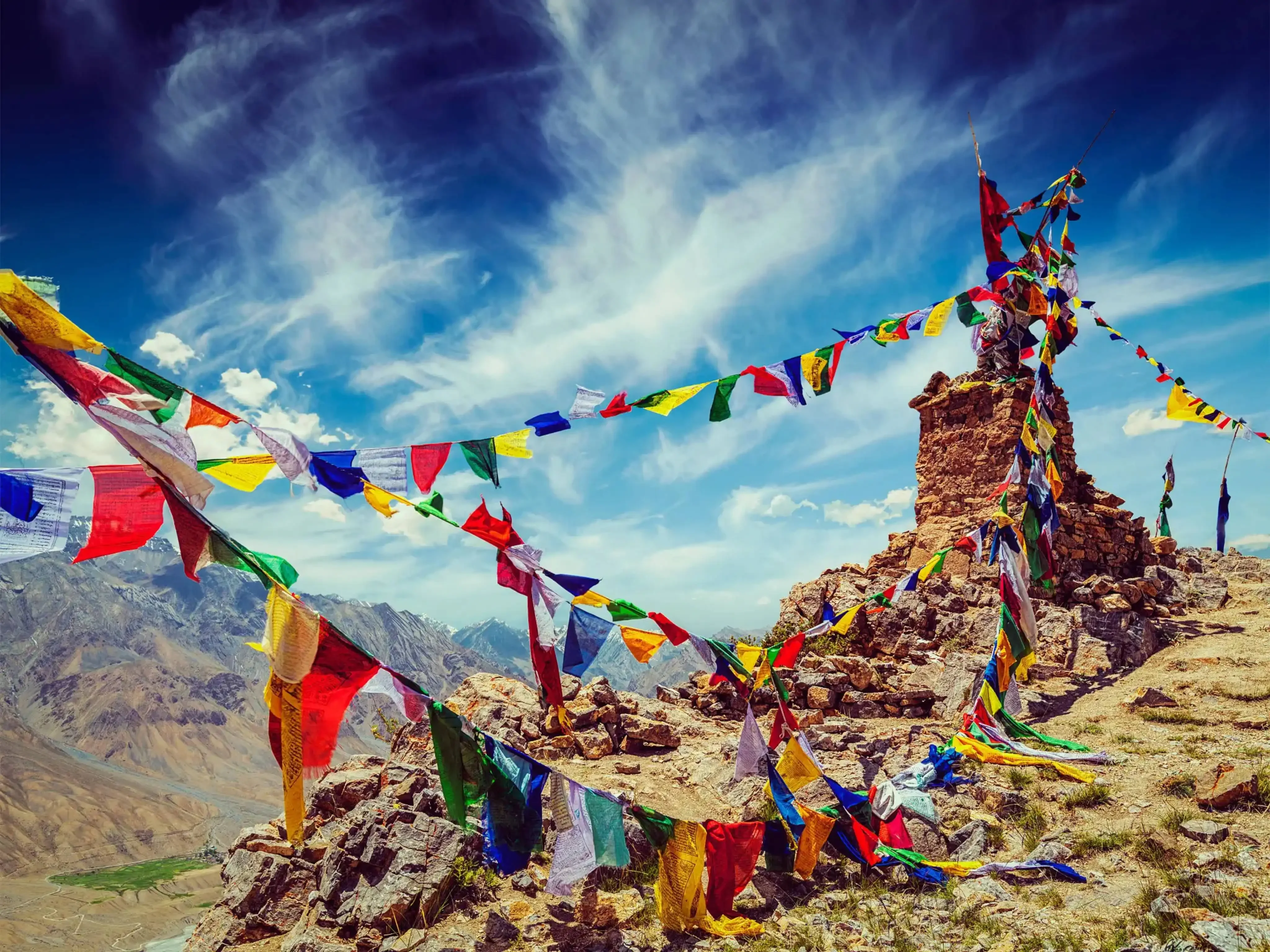 Colorful Tibetan prayer flags fluttering on a rocky mountain pass in the Himalayas, set against a dramatic blue sky with wispy clouds.
