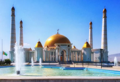 Turkmenbashi Ruhy Mosque in Gypjak, Turkmenistan, featuring a grand golden dome, four towering white minarets, and fountains in the foreground under a clear blue sky.
