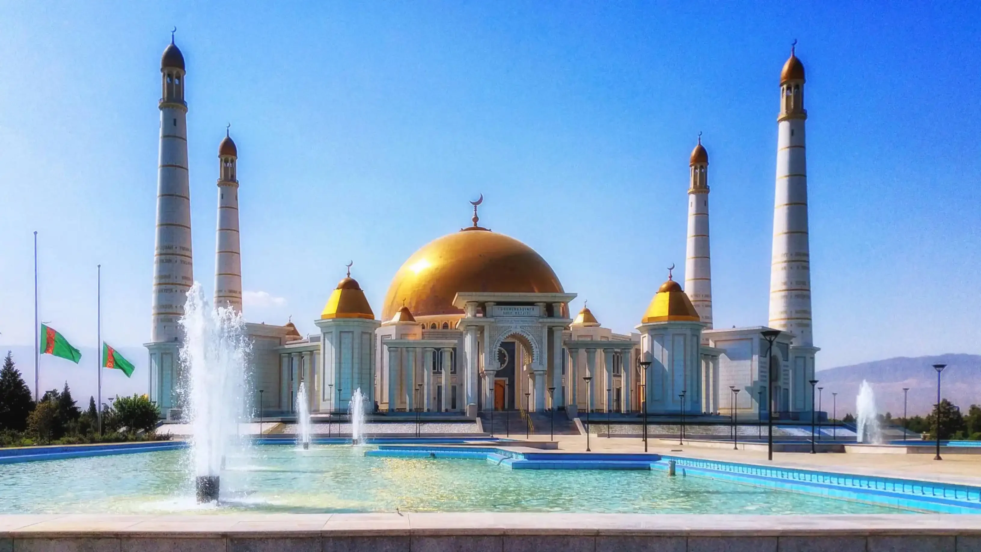 Turkmenbashi Ruhy Mosque in Gypjak, Turkmenistan, featuring a grand golden dome, four towering white minarets, and fountains in the foreground under a clear blue sky.