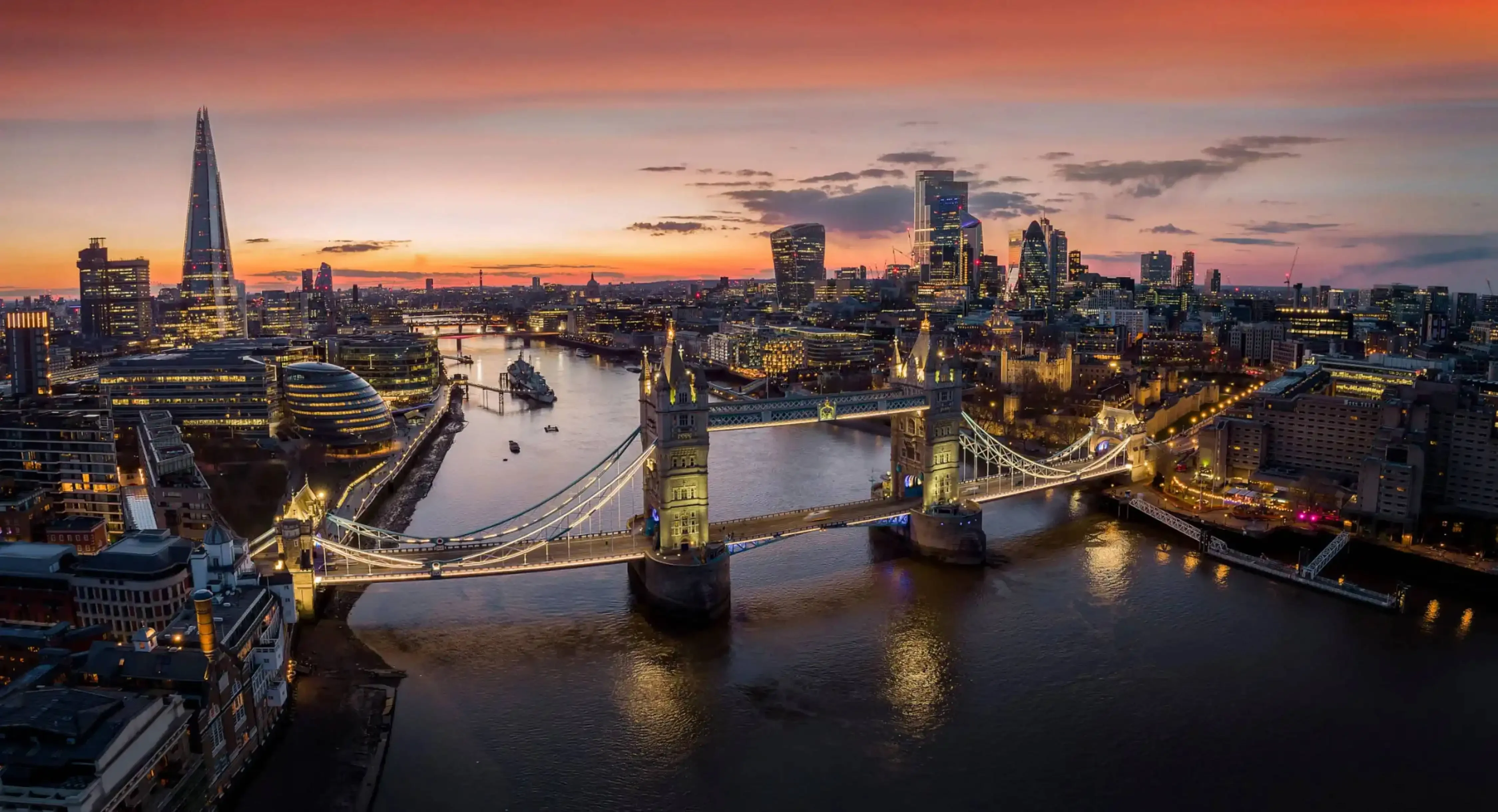 Sunset view over London with Tower Bridge illuminated and the Shard skyscraper in the background, reflecting on the River Thames.