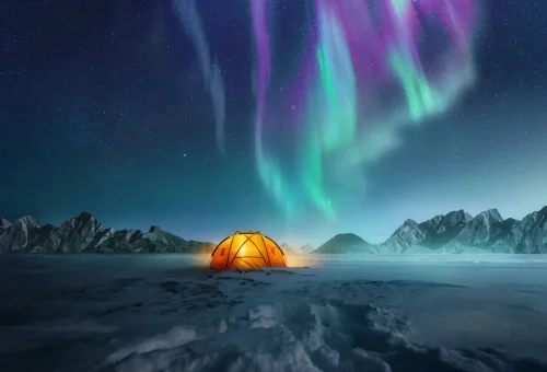 Glowing orange tent in a snowy Arctic landscape under a vivid aurora borealis with green and purple lights dancing across the night sky.