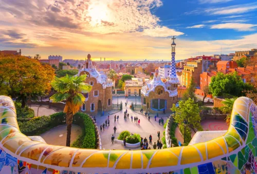 Colorful mosaic benches and whimsical architecture in Park Güell, Barcelona, with a vibrant sunset sky and cityscape in the background.
