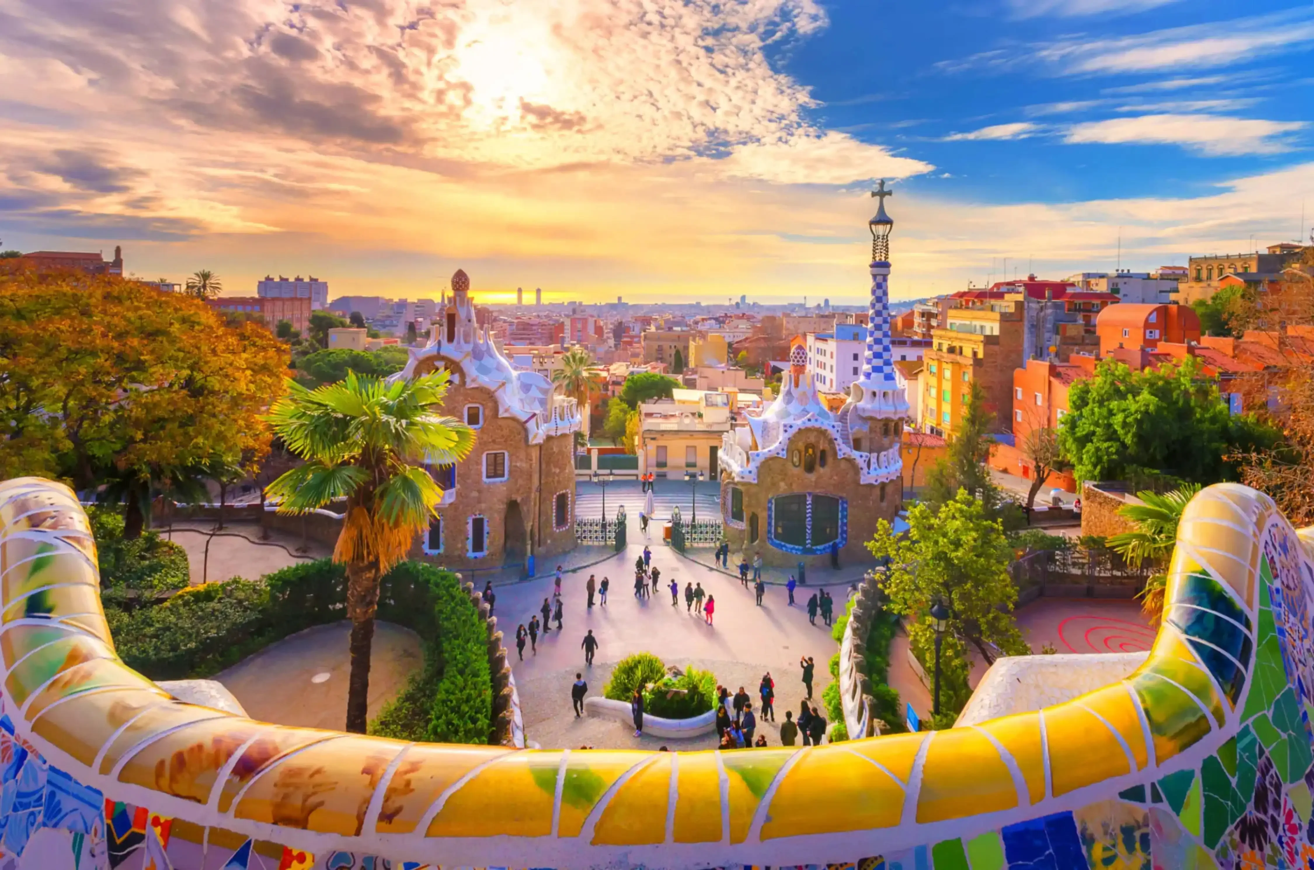 Colorful mosaic benches and whimsical architecture in Park Güell, Barcelona, with a vibrant sunset sky and cityscape in the background.