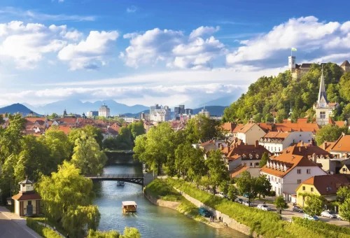 Scenic view of Ljubljana, Slovenia, featuring the Ljubljanica River, a charming bridge, red-roofed old town buildings, and Ljubljana Castle atop a lush green hill under a bright sky.