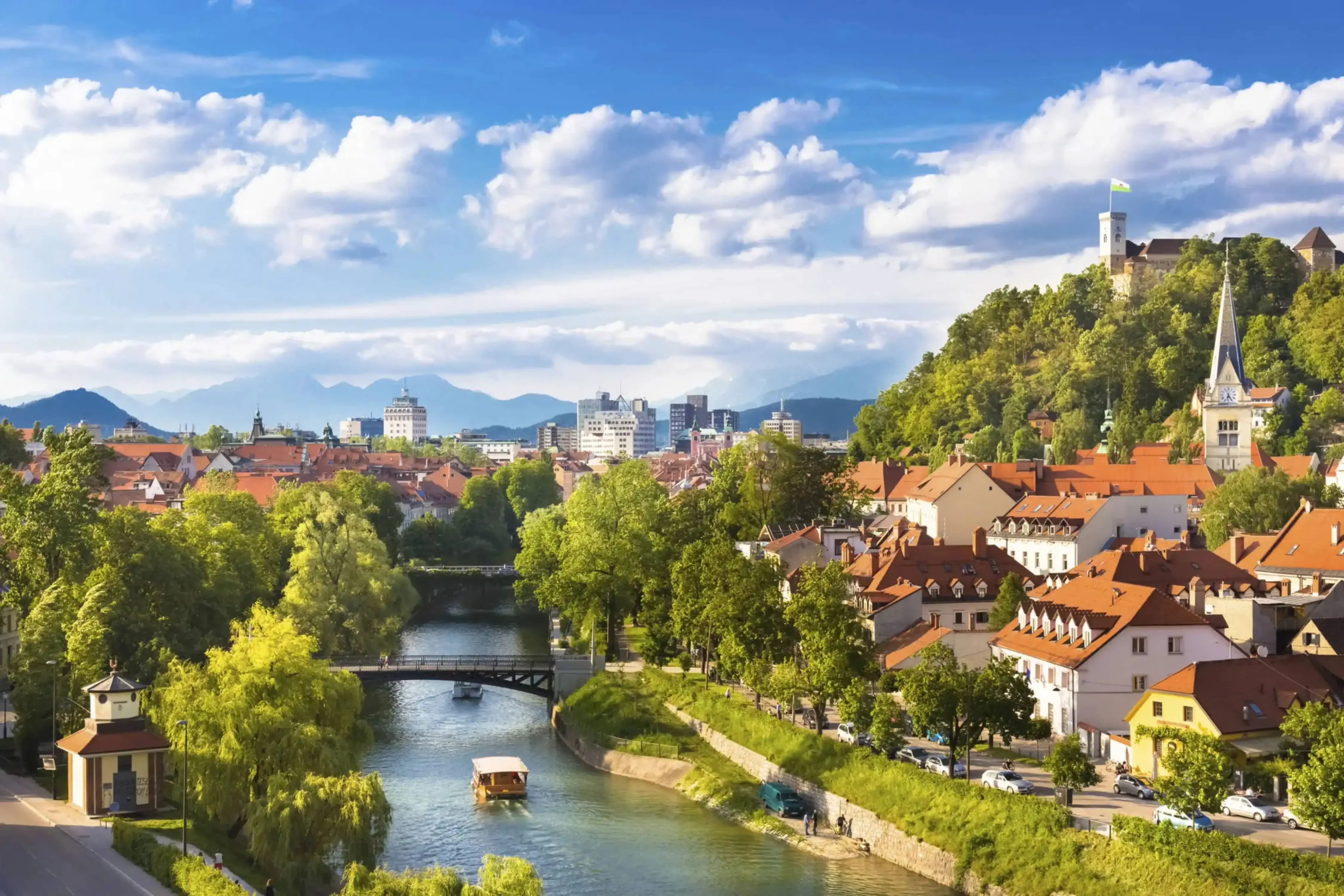 Scenic view of Ljubljana, Slovenia, featuring the Ljubljanica River, a charming bridge, red-roofed old town buildings, and Ljubljana Castle atop a lush green hill under a bright sky.