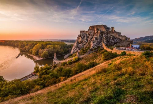 Aerial view of Devín Castle in Slovakia at sunset, perched on a rocky cliff above the confluence of the Danube and Morava rivers, with rolling hills and golden light in the background.