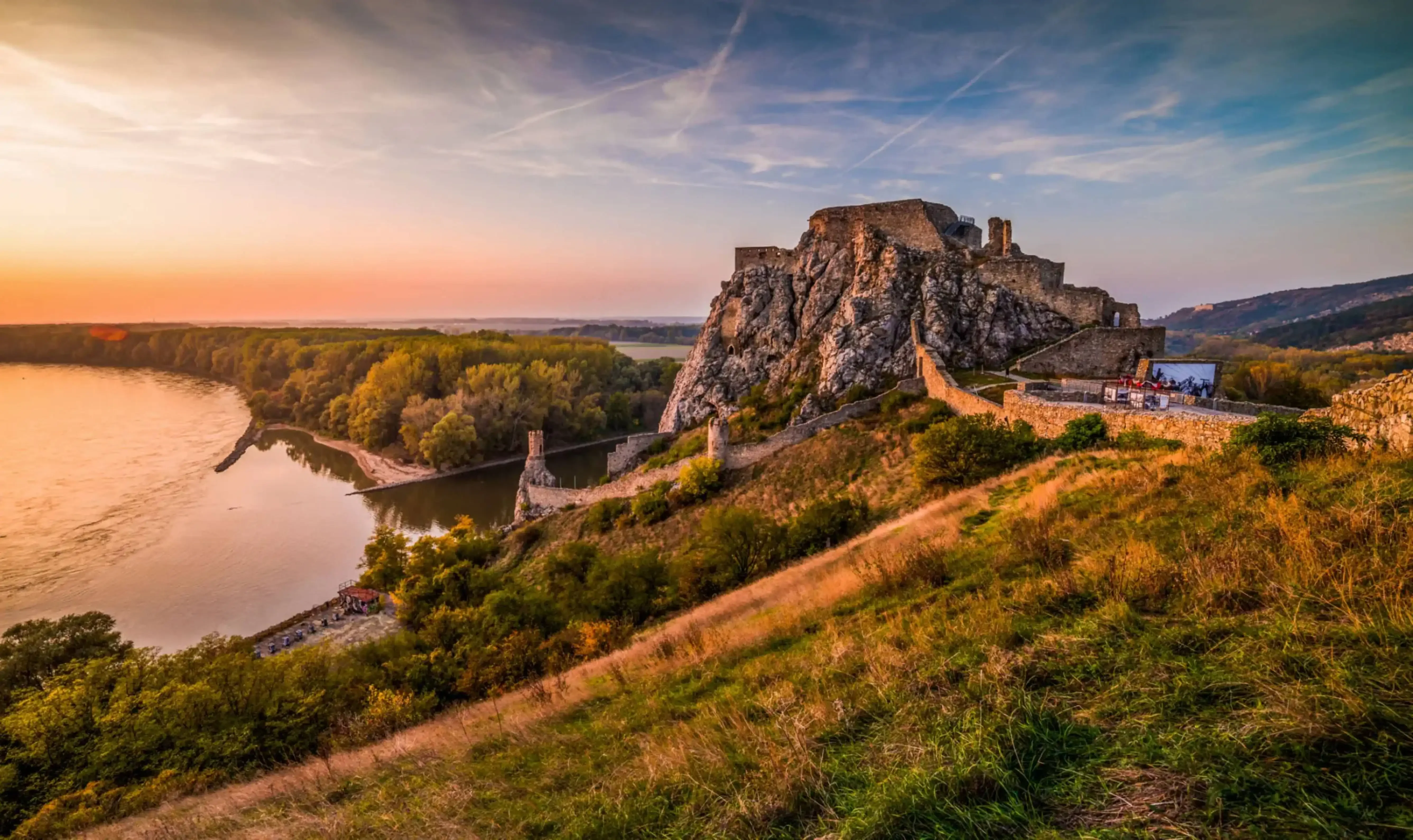 Aerial view of Devín Castle in Slovakia at sunset, perched on a rocky cliff above the confluence of the Danube and Morava rivers, with rolling hills and golden light in the background.