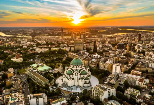Sunset aerial view of Belgrade, Serbia, featuring the Church of Saint Sava with its prominent green dome surrounded by urban cityscape and glowing sky.