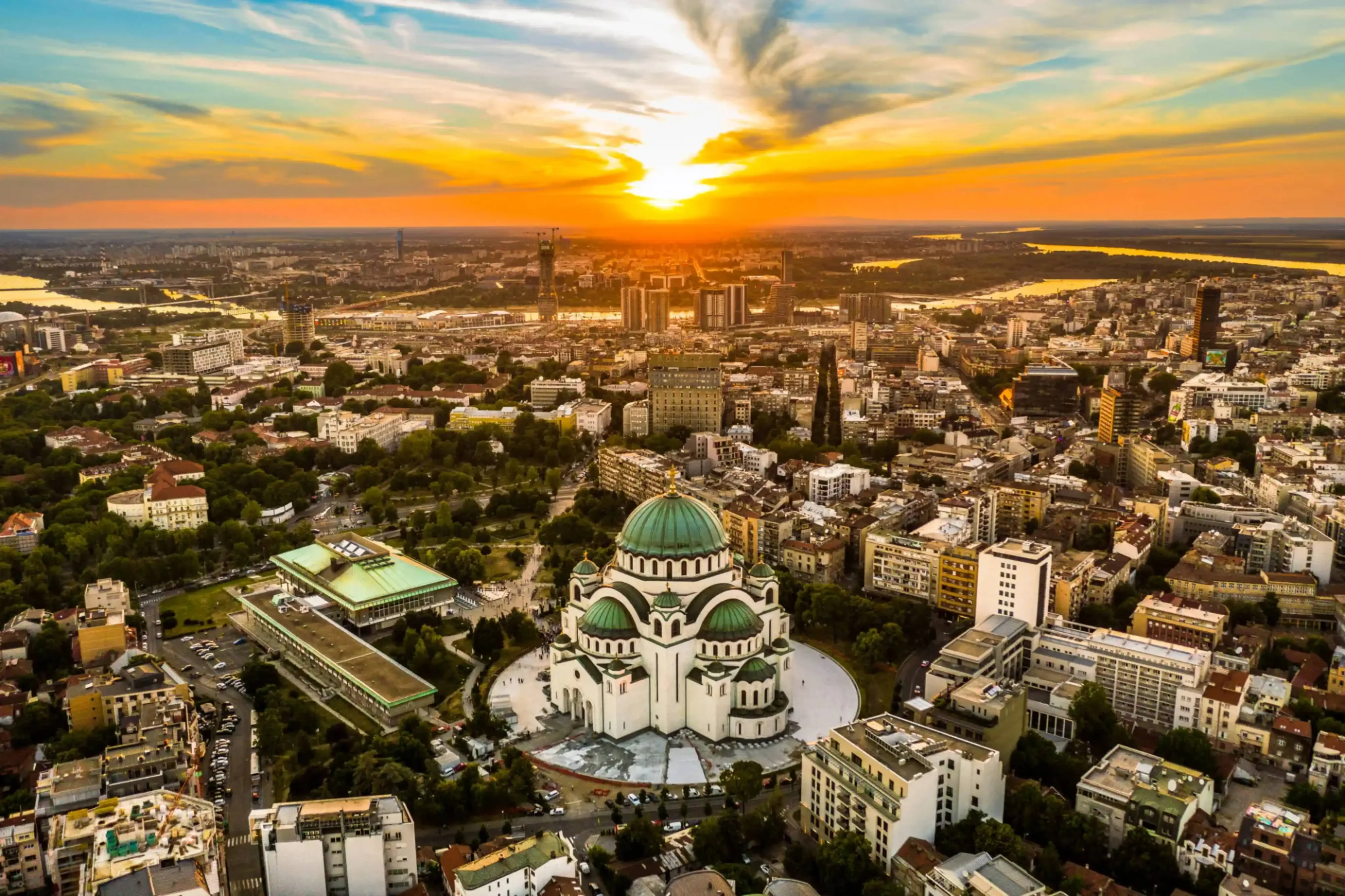 Sunset aerial view of Belgrade, Serbia, featuring the Church of Saint Sava with its prominent green dome surrounded by urban cityscape and glowing sky.