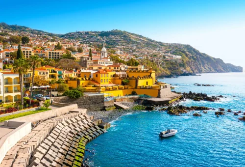 Scenic coastal view of Funchal, the capital city of Madeira, Portugal, with colorful buildings, terraced hillsides, and a calm blue sea along a stone promenade.