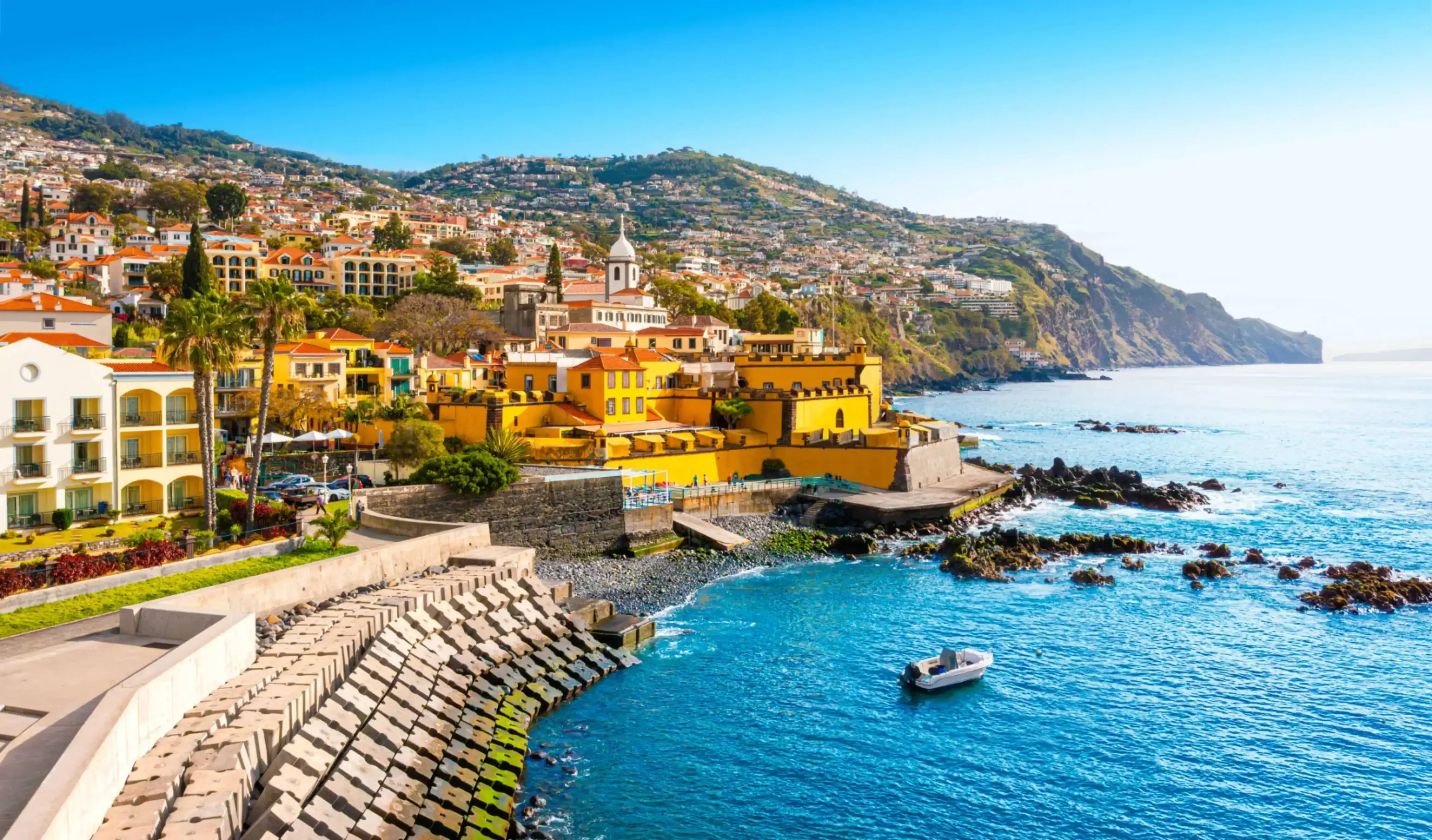 Scenic coastal view of Funchal, the capital city of Madeira, Portugal, with colorful buildings, terraced hillsides, and a calm blue sea along a stone promenade.