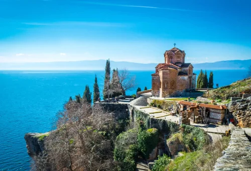 Scenic view of the Church of St. John at Kaneo perched on a cliff above Lake Ohrid in North Macedonia, surrounded by trees and overlooking the tranquil blue water.