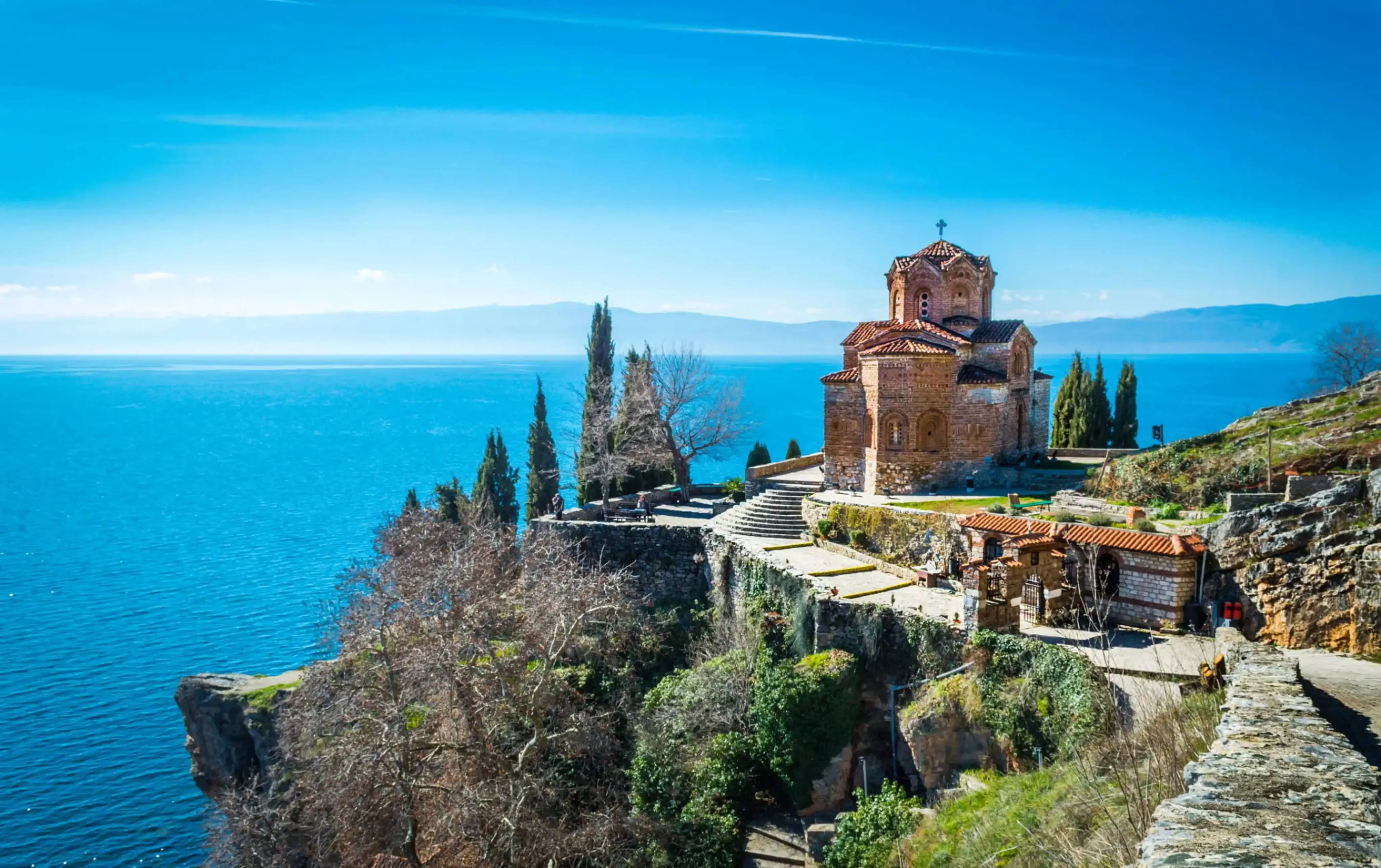 Scenic view of the Church of St. John at Kaneo perched on a cliff above Lake Ohrid in North Macedonia, surrounded by trees and overlooking the tranquil blue water.