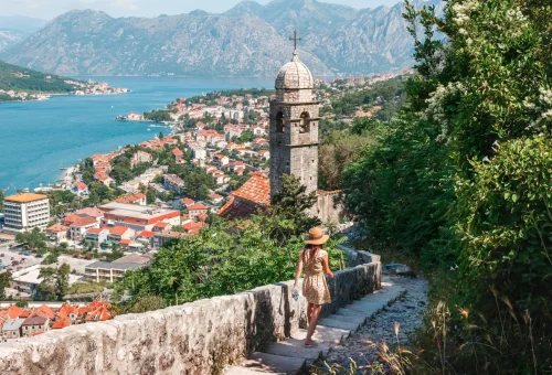 Woman walking down a stone path toward the Bay of Kotor, with a historic bell tower and red-roofed town nestled along Montenegro's Adriatic coastline.